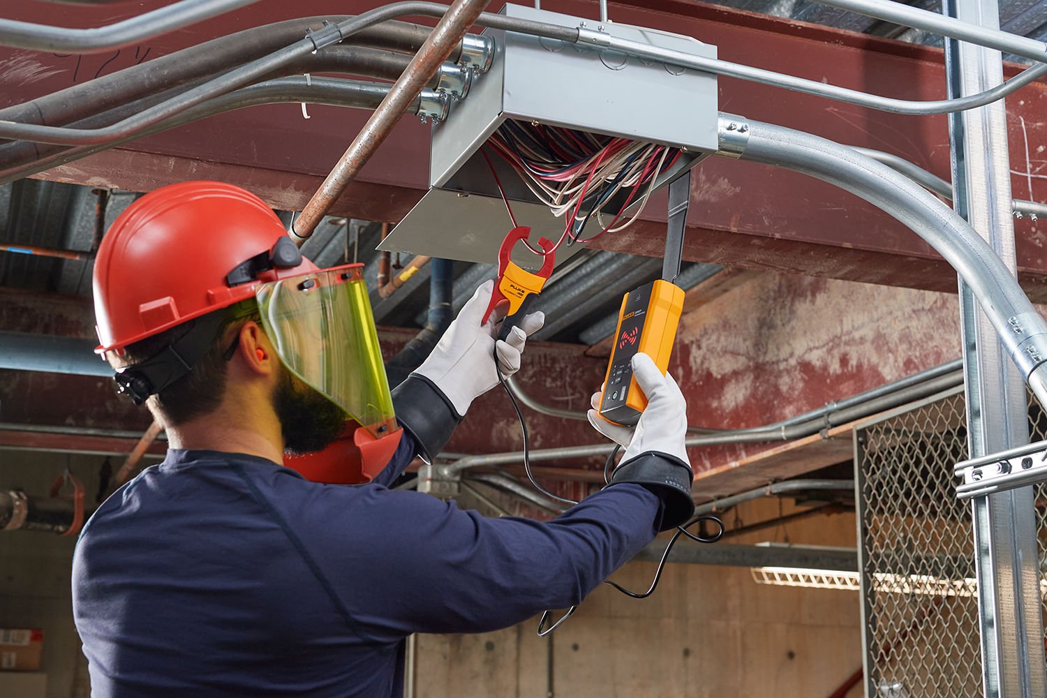Electrician in face shield and gloves uses a current clamp and wire tracer transmitter on wiring in a junction box