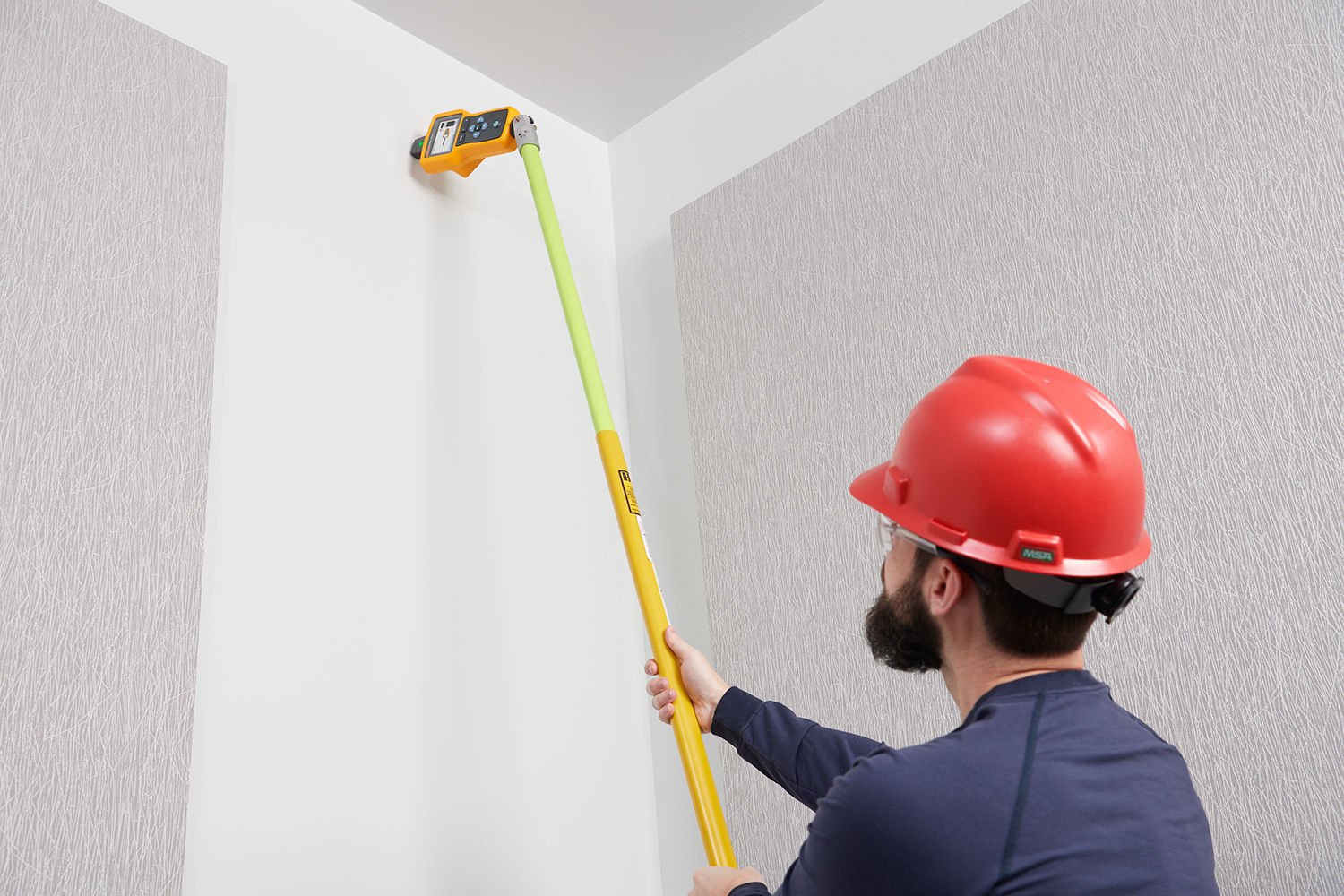 En man med en röd skyddshjälm använder en kabelsökare i änden av en lång stav för att hitta kablar högt upp i en vägg