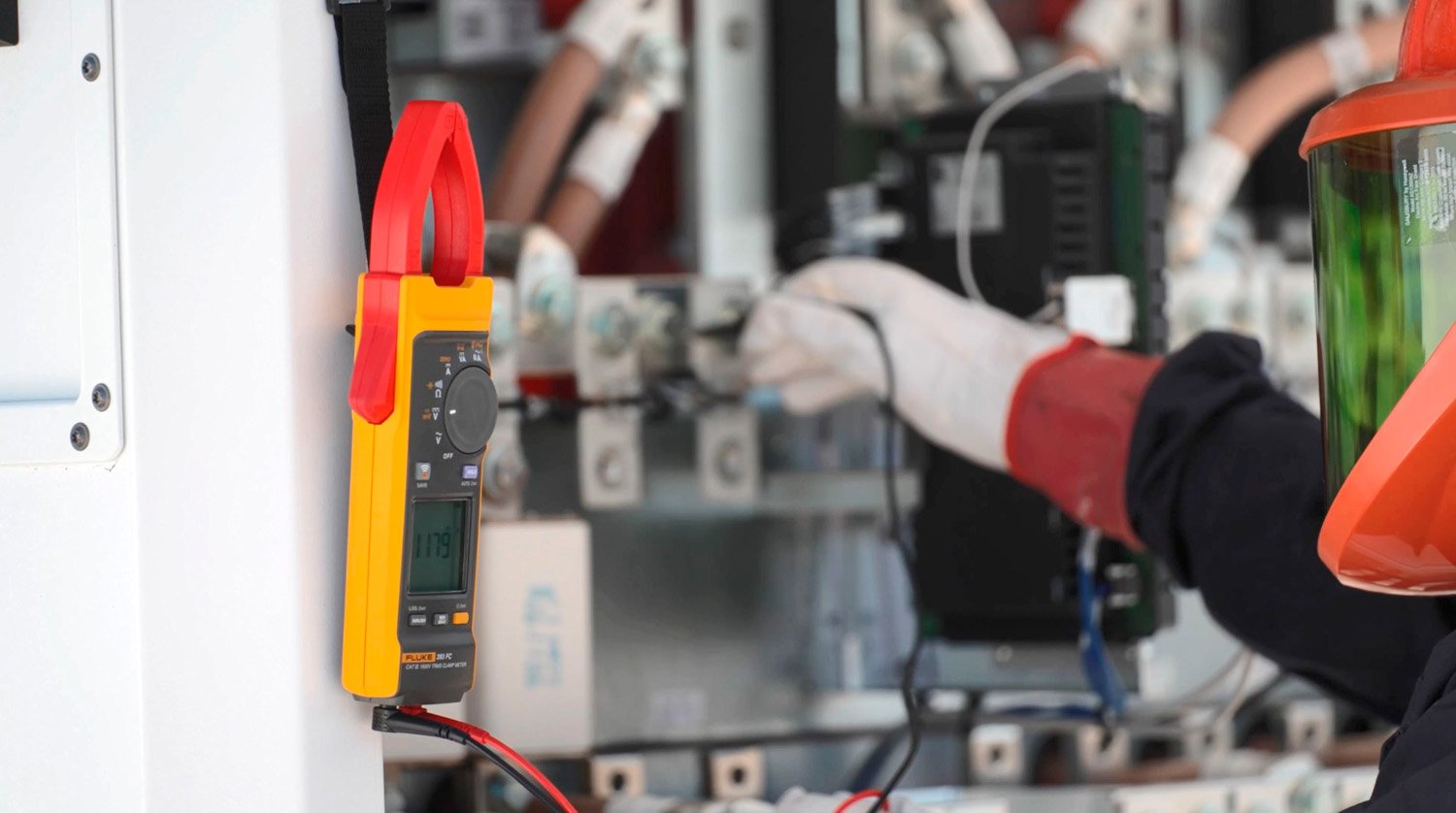 Eine Fluke 393 FC Strommesszange hängt neben einem geöffneten Wechselrichter, während ein Techniker, der PSA (inklusive Handschuhe und Gesichtsschutz) trägt, mit den Messleitungen eine Messung vornimmt.