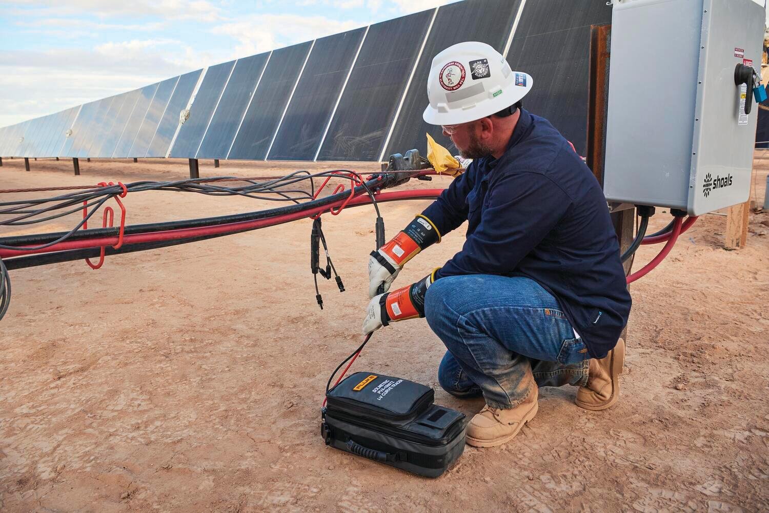 Technician kneeling beside solar PV array, connecting a tool to wiring