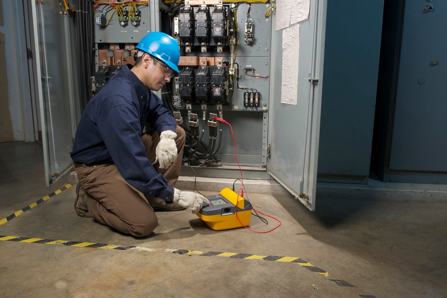 An electrician kneels before an open electrical panel, making measurements with an insulation tester